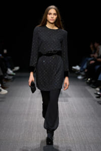 Model on a fashion runway wearing a black dotted long-sleeve top tucked into a matching dotted tunic and loose black pants, holding a small black clutch.