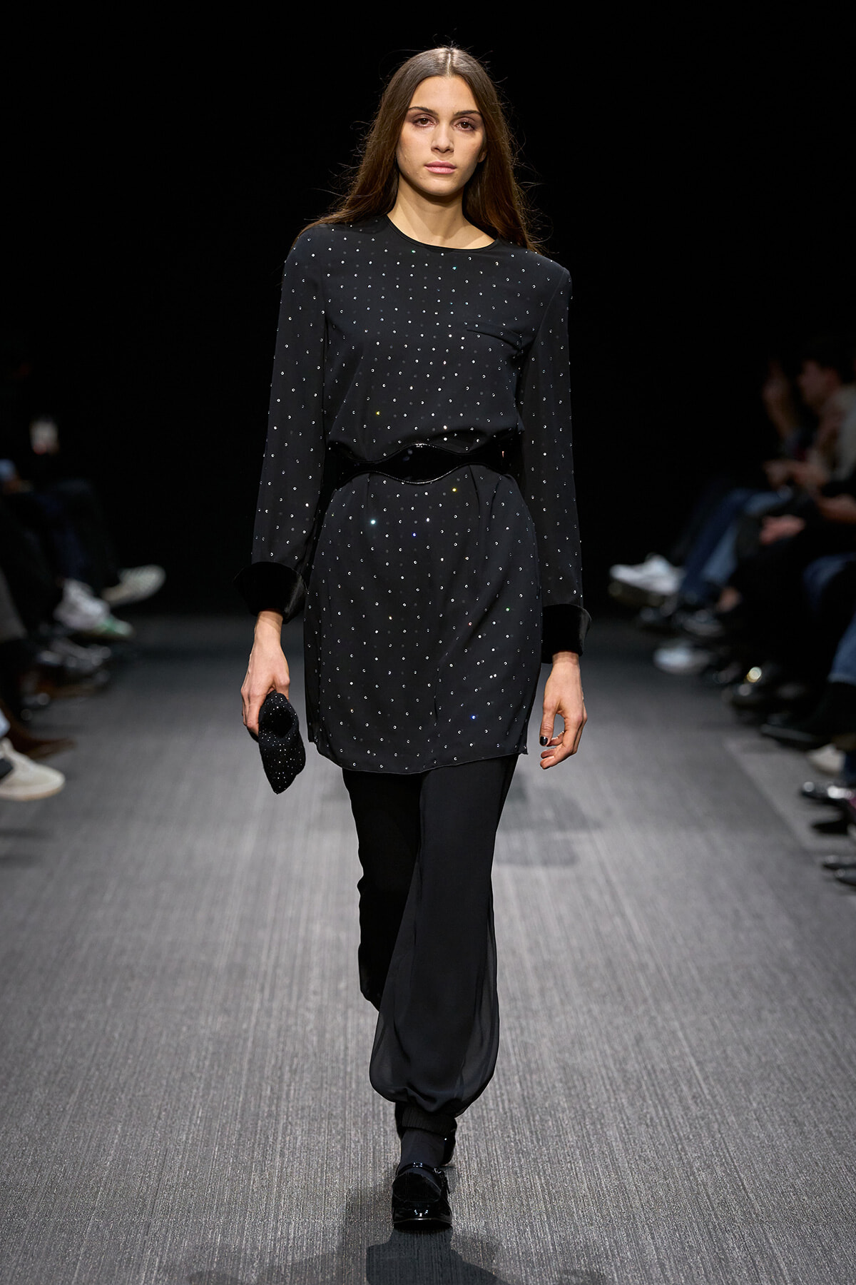 Model on a fashion runway wearing a black dotted long-sleeve top tucked into a matching dotted tunic and loose black pants, holding a small black clutch.