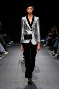 Model on the runway wearing a metallic silver blazer with black lapels, black pants, a black beret, and a small black crossbody bag