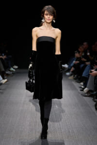 Model on a runway wearing a strapless black velvet dress, long black gloves, crystal earrings, and a black handbag.
