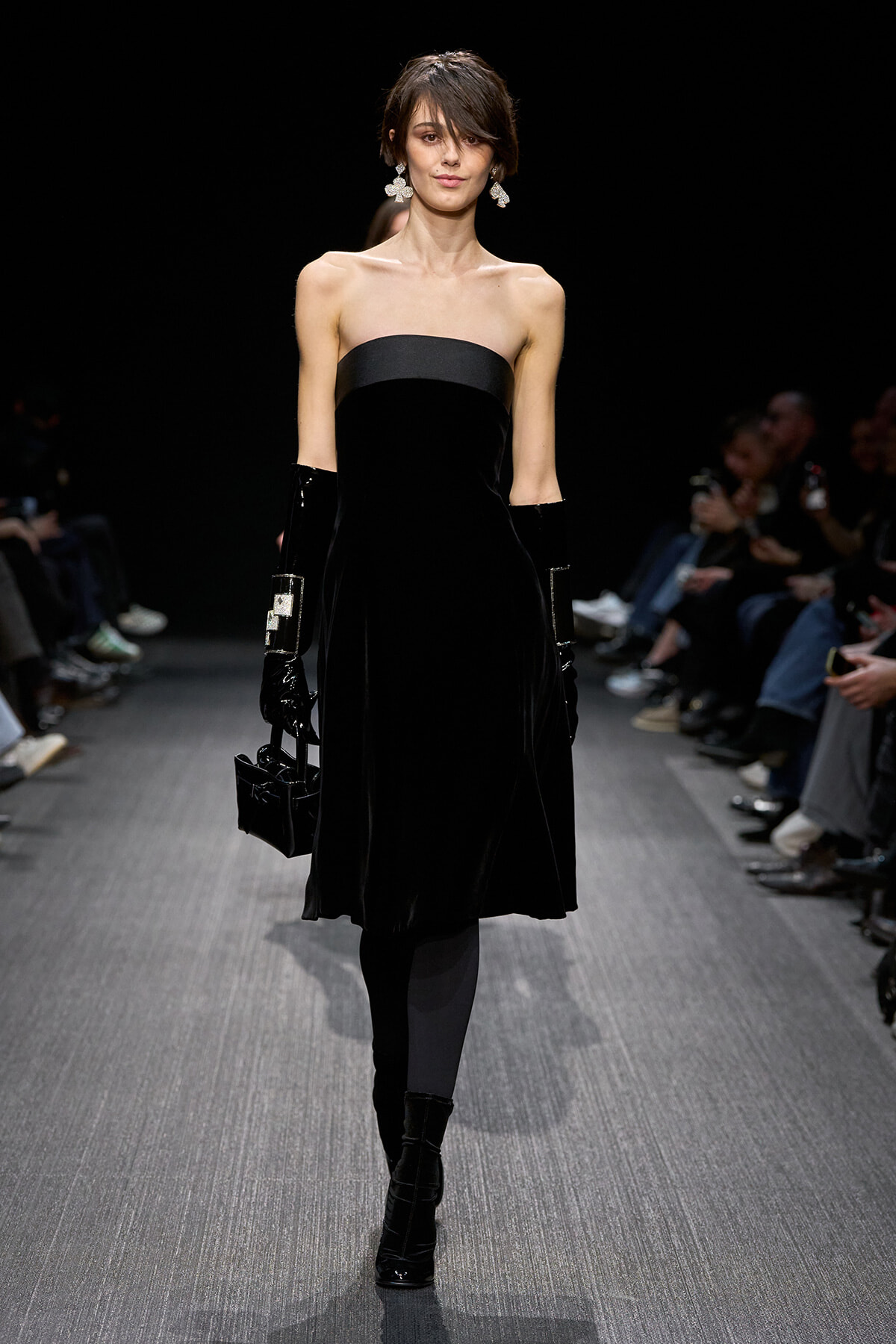 Model on a runway wearing a strapless black velvet dress, long black gloves, crystal earrings, and a black handbag.