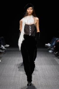 Model walking a runway in black velvet pants, a sheer mesh top with white fringe scarf, and a black beret.