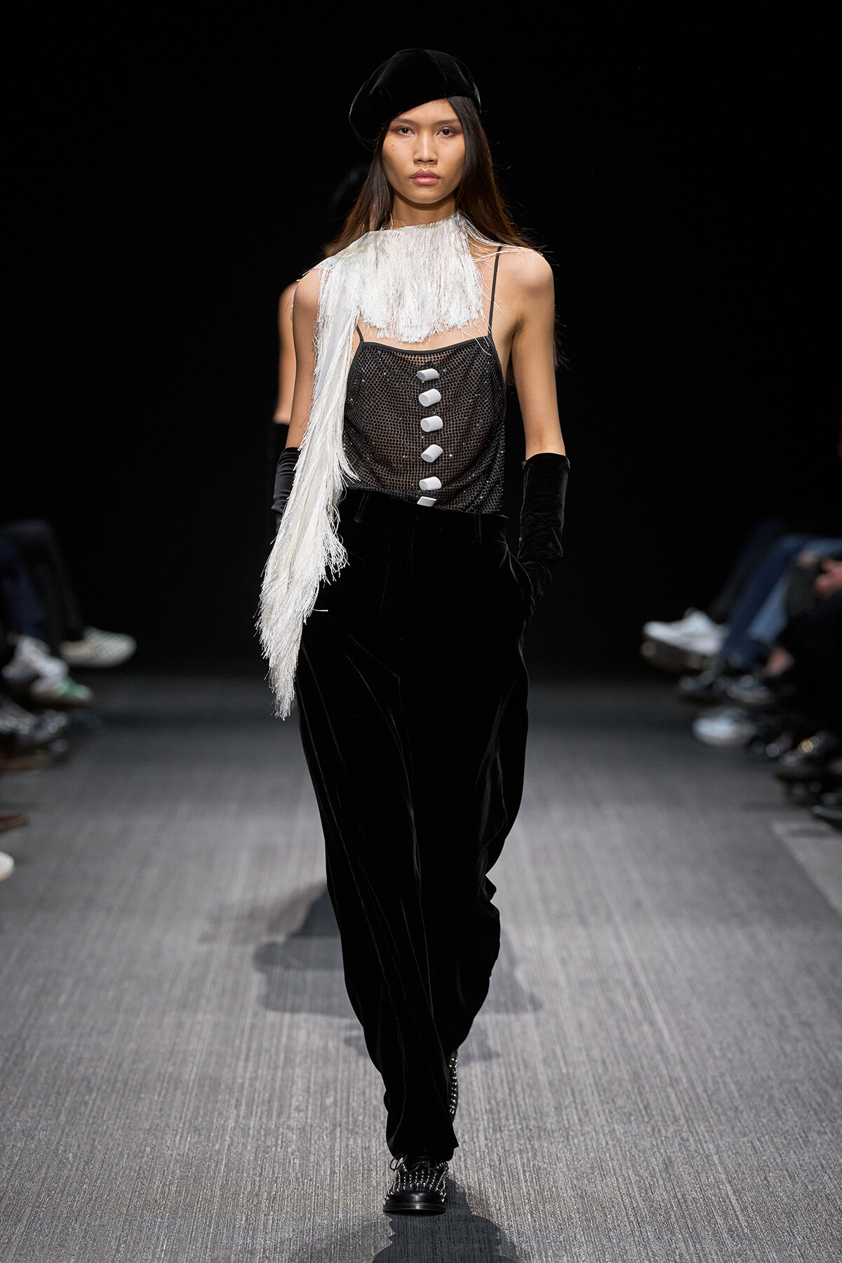 Model walking a runway in black velvet pants, a sheer mesh top with white fringe scarf, and a black beret.
