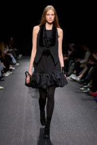 Model on a runway wearing an all-black ensemble: sleeveless vest, ruffled mini skirt, black tights, studded ankle boots, layered necklace, and a small black handbag.”