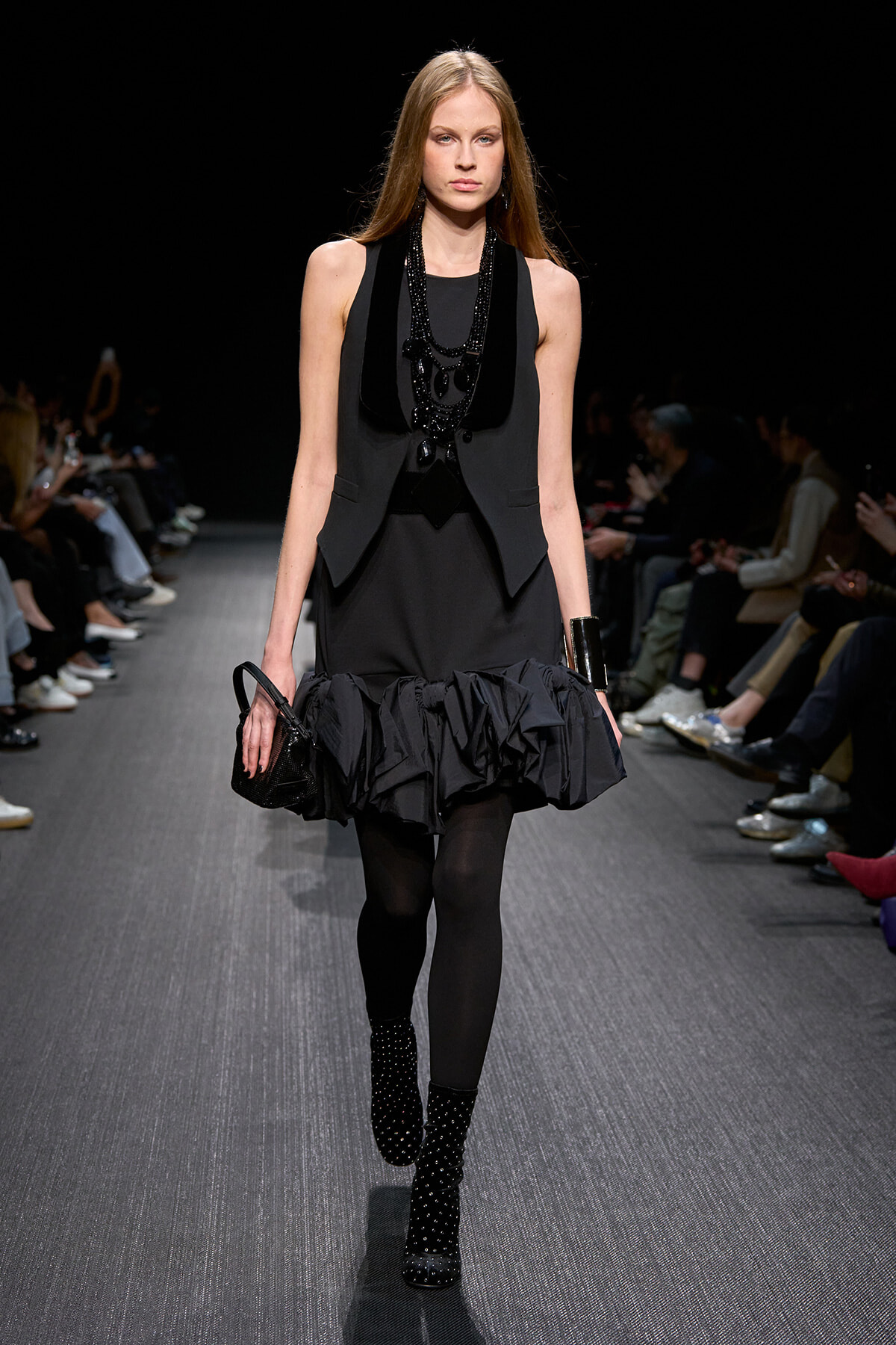 Model on a runway wearing an all-black ensemble: sleeveless vest, ruffled mini skirt, black tights, studded ankle boots, layered necklace, and a small black handbag.”