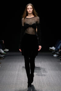 Runway model in an all-black outfit: sheer mesh top with glittery sleeves, a black velvet skirt, and tall boots.