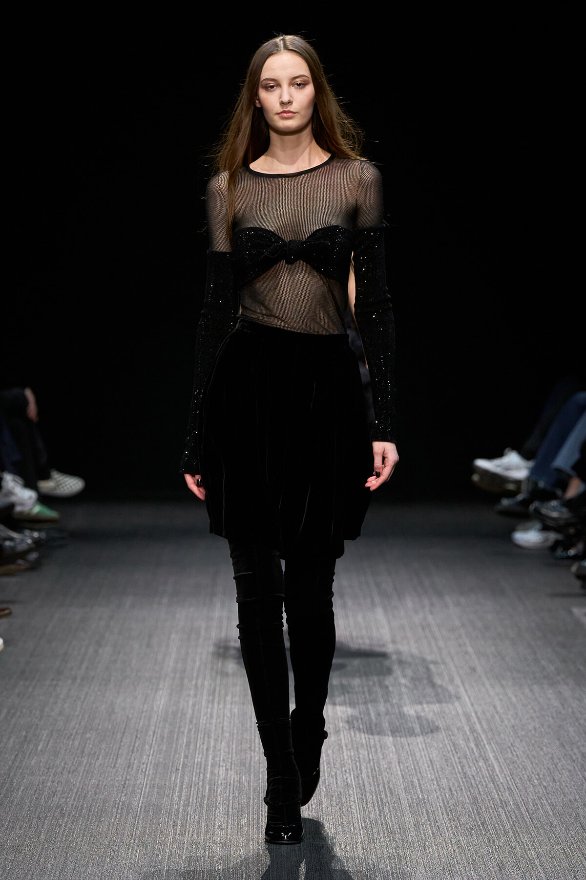Runway model in an all-black outfit: sheer mesh top with glittery sleeves, a black velvet skirt, and tall boots.