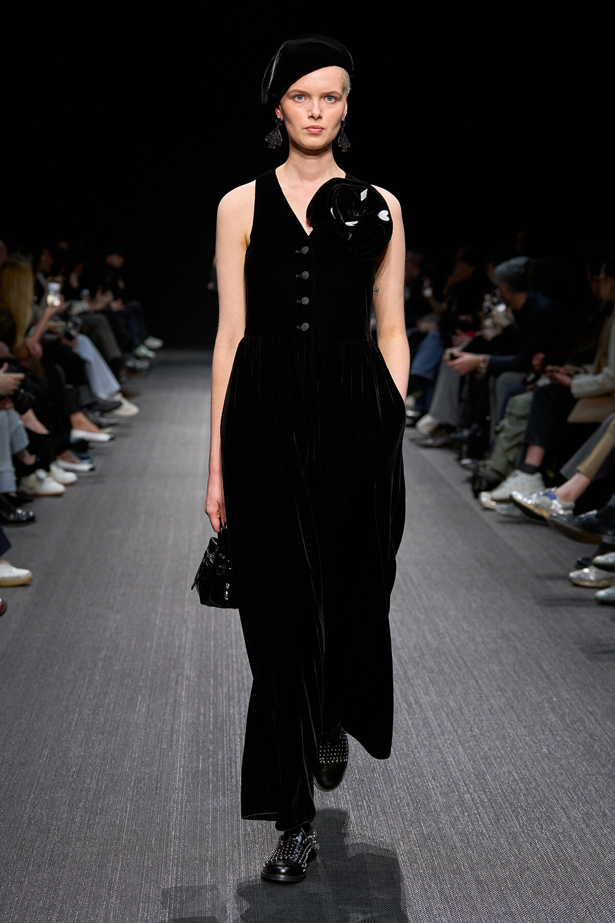 Model walks a runway in a black velvet sleeveless gown with front buttons and a large fabric flower on the shoulder, wearing a black beret.