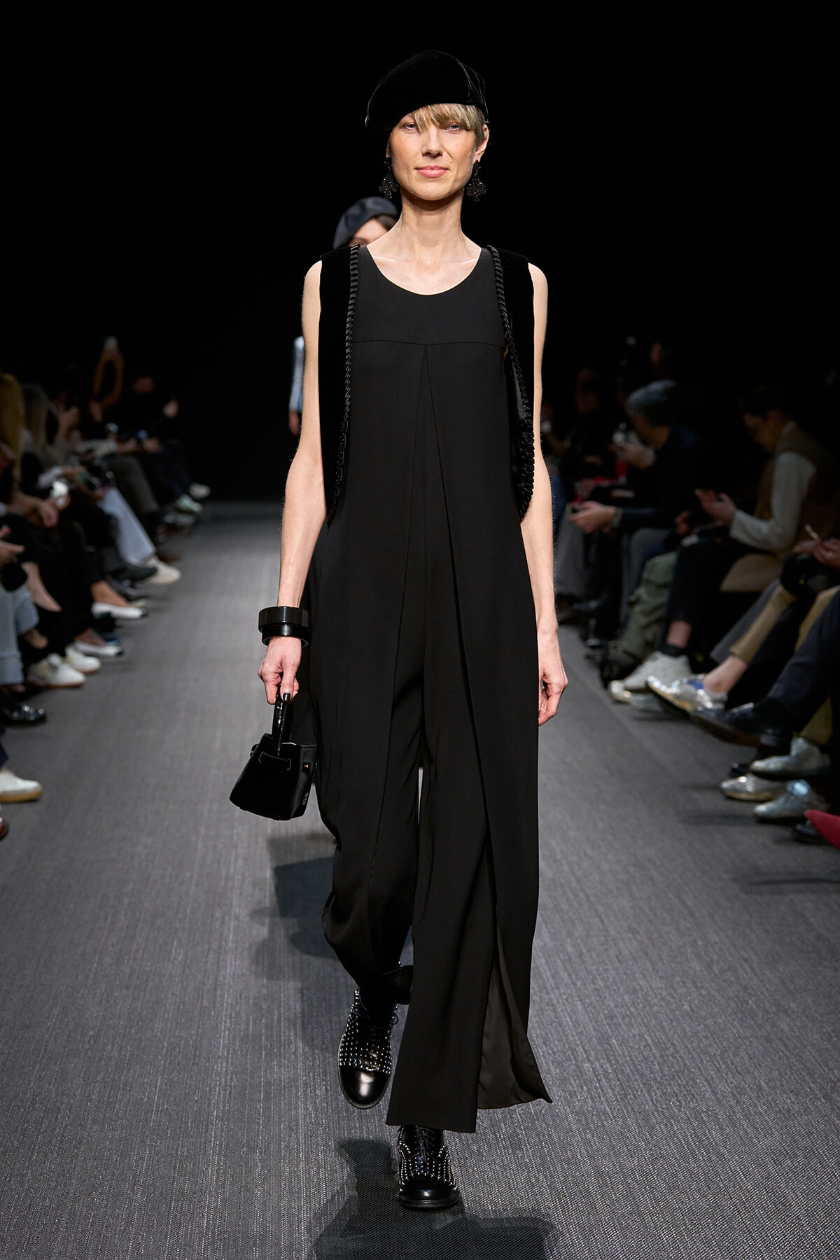 Model walks the runway in an all-black sleeveless jumpsuit with draped panels, black hat, and studded boots, carrying a small black handbag.