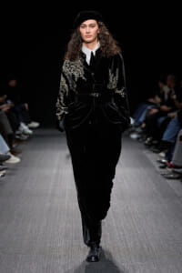 Model walks the runway in a black velvet suit with gold embroidery, white shirt, belt, and beret; audience visible in the background.
