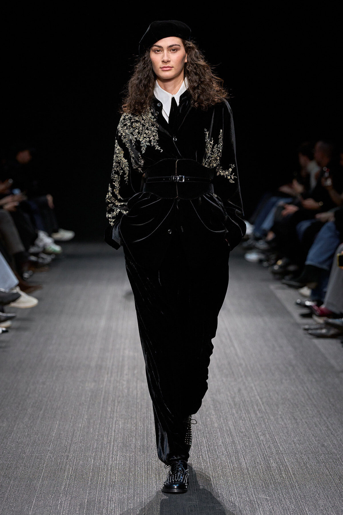 Model walks the runway in a black velvet suit with gold embroidery, white shirt, belt, and beret; audience visible in the background.