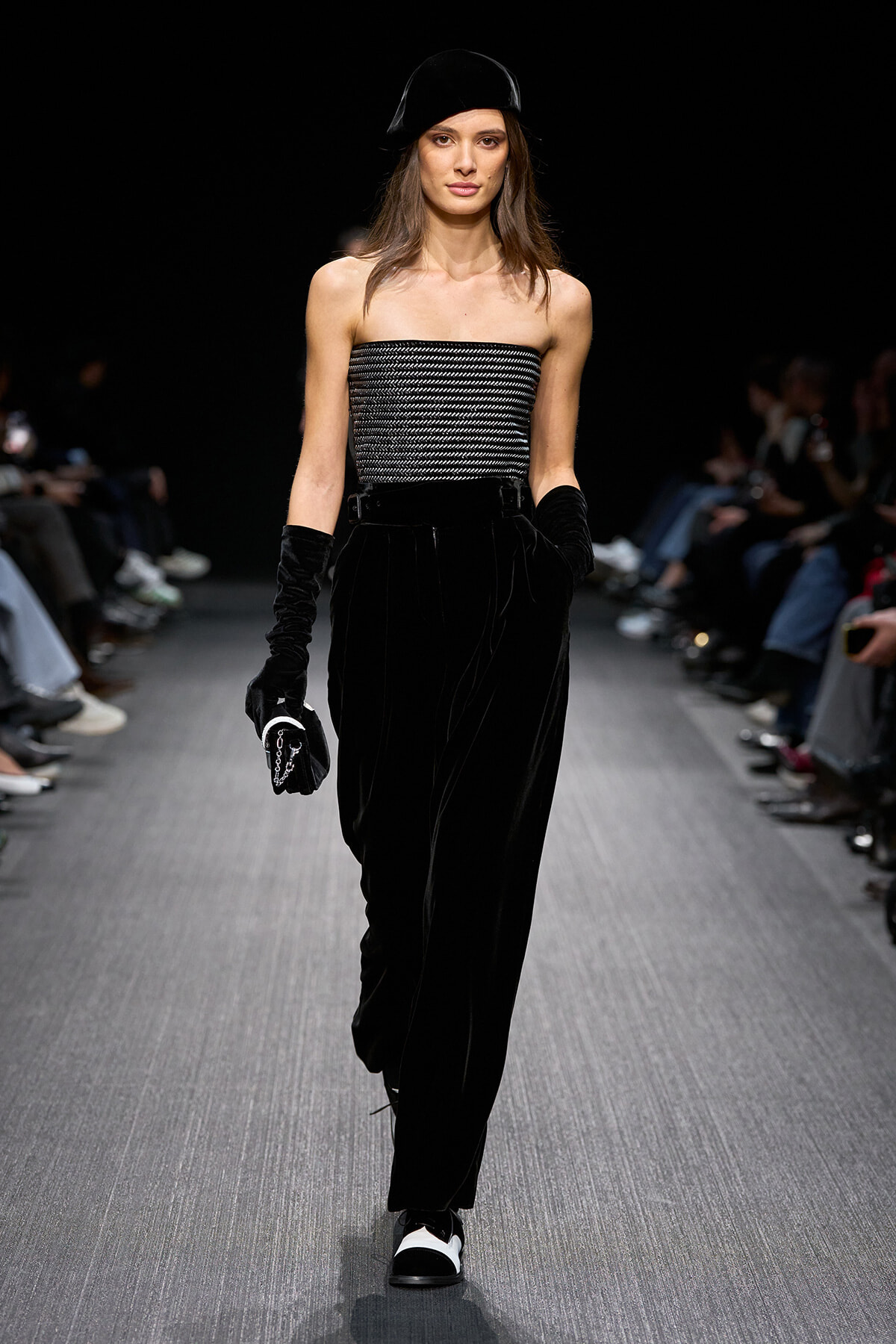 Model walking a fashion runway in a black velvet jumpsuit-like ensemble with a striped strapless top and matching velvet gloves, topped with a black beret on a dark stage.