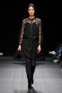 Model on a dim runway wearing a black sequined dress with sheer lattice sleeves and a zip-front vest, carrying a small black handbag.