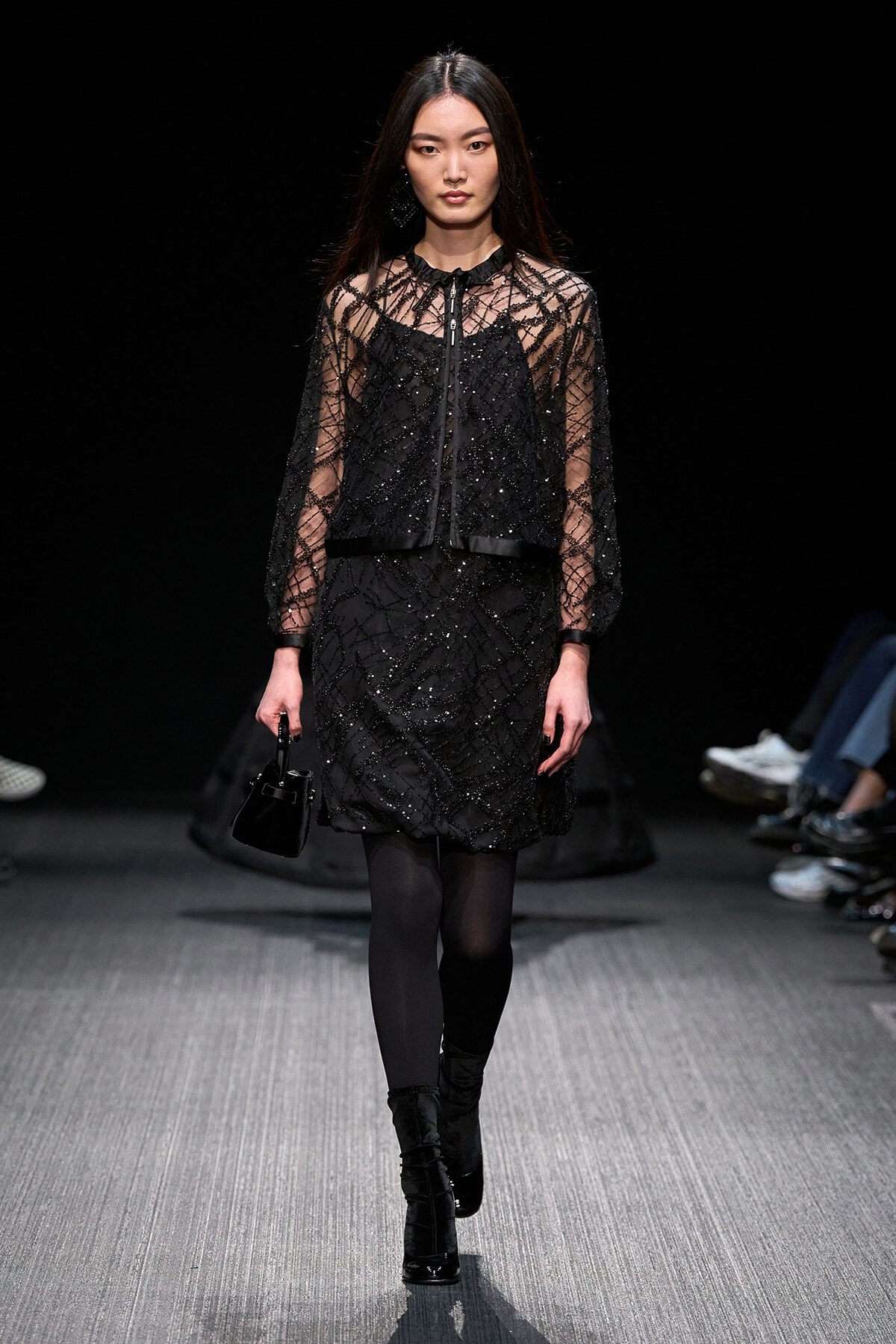 Model on a dim runway wearing a black sequined dress with sheer lattice sleeves and a zip-front vest, carrying a small black handbag.