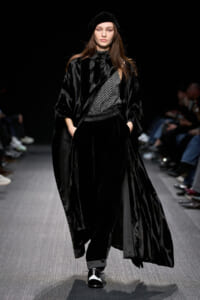 Model on a runway wearing an all-black velvet ensemble with a flowing cape, patterned top, and beret.