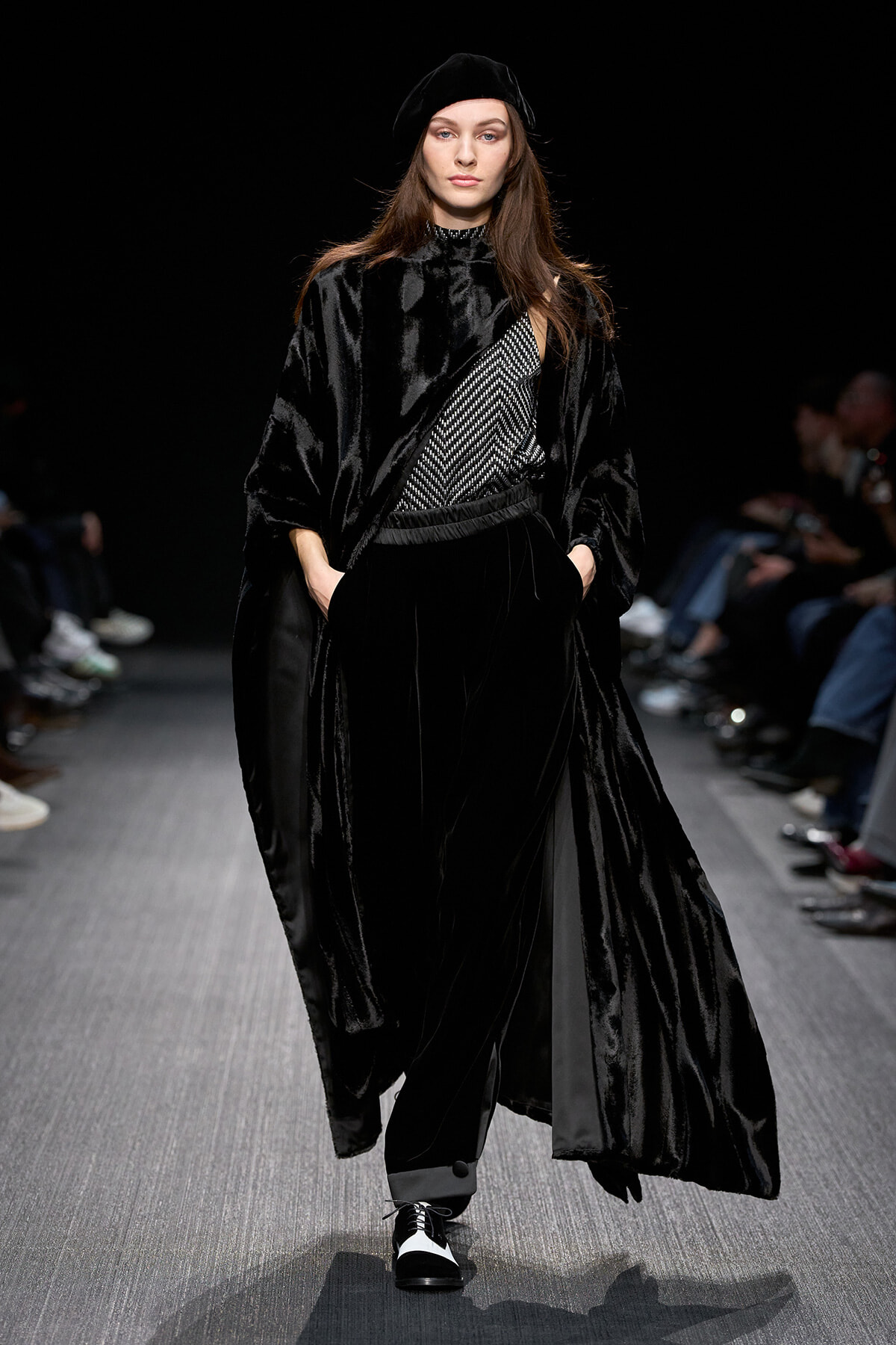 Model on a runway wearing an all-black velvet ensemble with a flowing cape, patterned top, and beret.