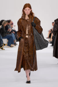 Model on a fashion runway wearing a brown snakeskin coat with belt, carrying a large gray snakeskin tote and gloves.