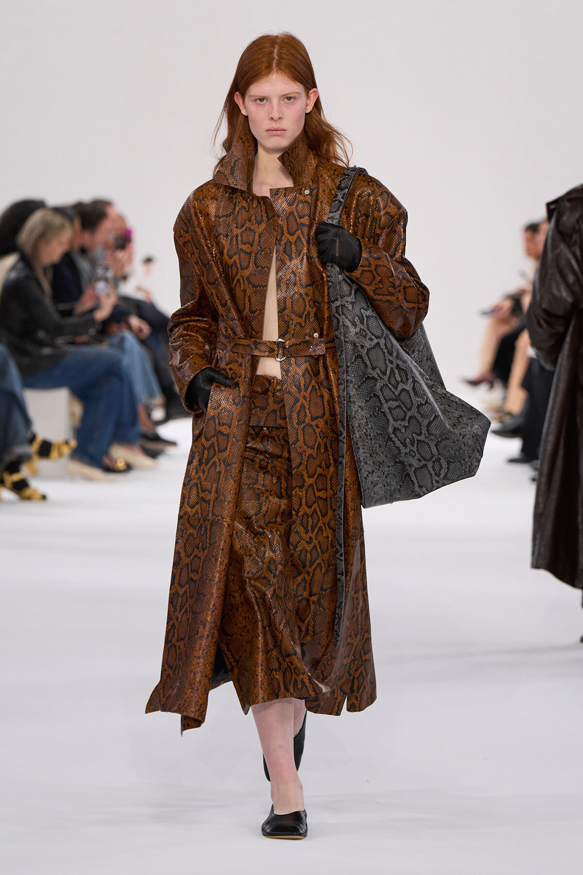 Model on a fashion runway wearing a brown snakeskin coat with belt, carrying a large gray snakeskin tote and gloves.