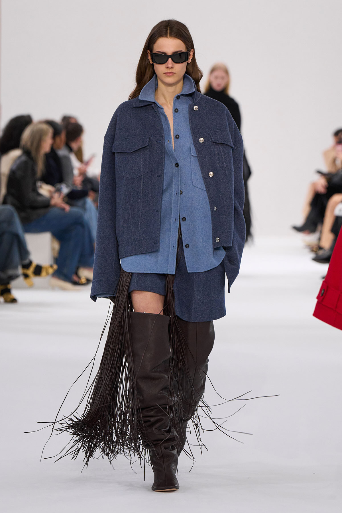 Runway model in a blue denim ensemble with oversized jacket, shirt, and shorts, plus knee-high black fringe boots and sunglasses.