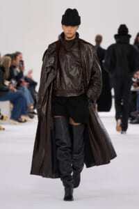 Model walks the runway in a shiny dark brown oversized coat, knee-high black boots, and a black knit hat; audience seated along the catwalk in the background.
