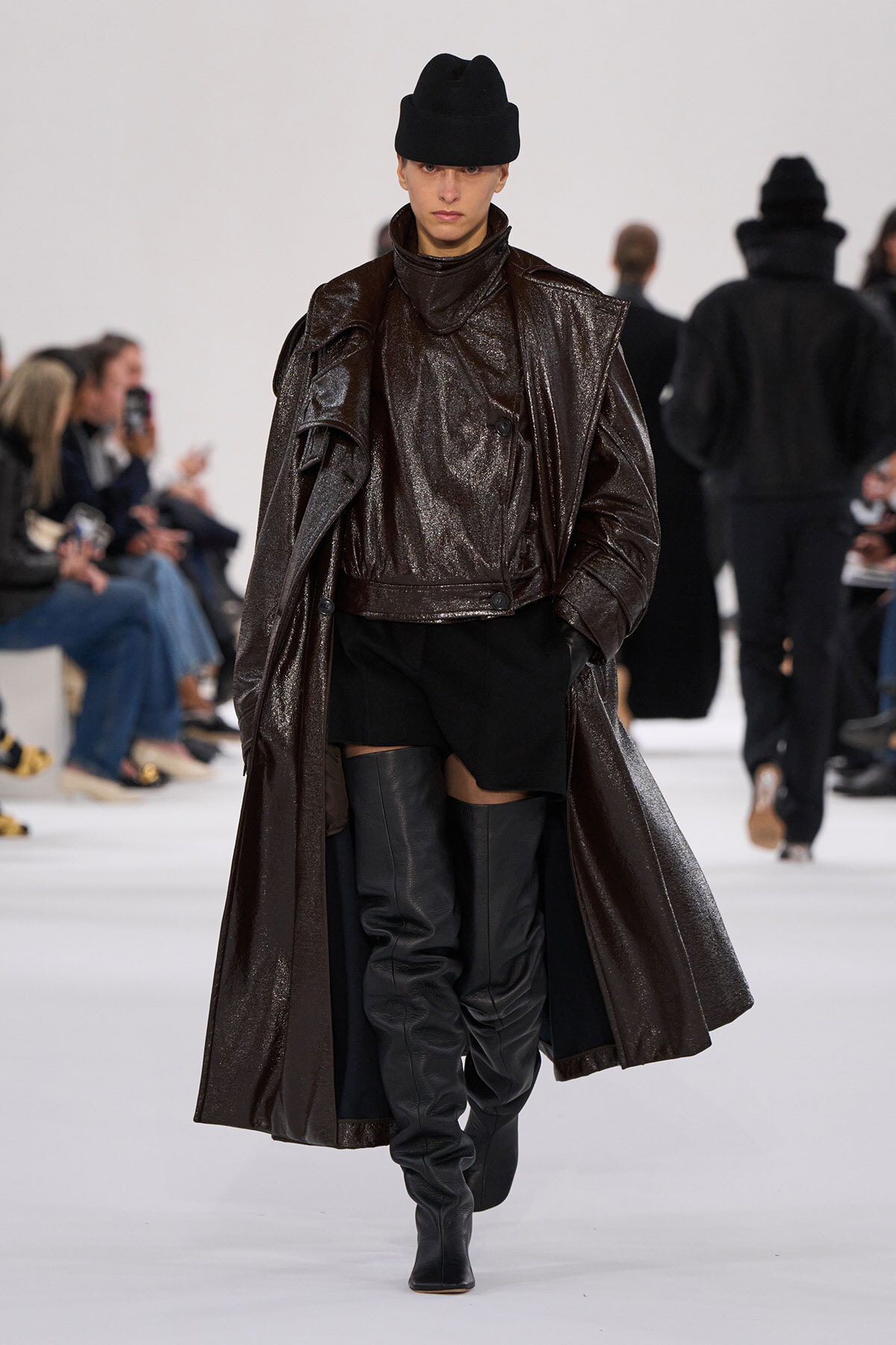 Model walks the runway in a shiny dark brown oversized coat, knee-high black boots, and a black knit hat; audience seated along the catwalk in the background.
