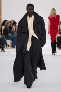 Model walks a fashion runway in an oversized dark pinstripe coat over a cream shirt and wide-leg trousers, hands in pockets.