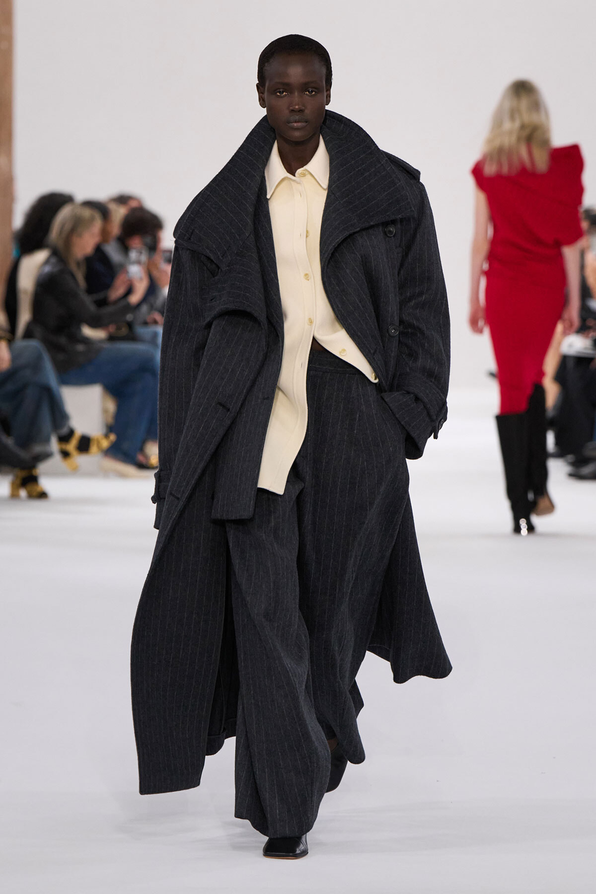 Model walks a fashion runway in an oversized dark pinstripe coat over a cream shirt and wide-leg trousers, hands in pockets.