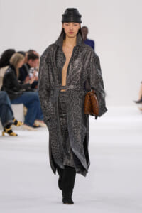 Model walking a runway in a gray snakeskin-pattern long coat, belt at the waist, black boots, and a black leather hat, holding a brown clutch.