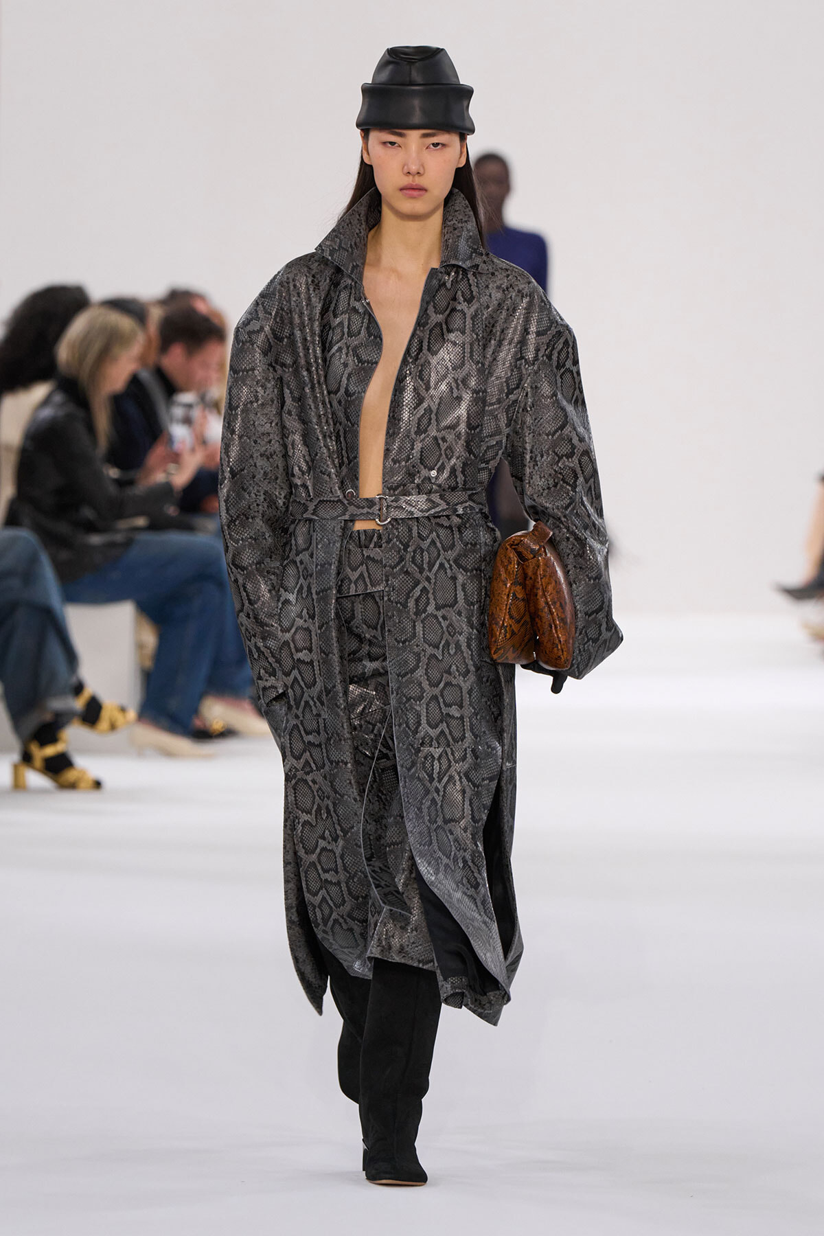 Model walking a runway in a gray snakeskin-pattern long coat, belt at the waist, black boots, and a black leather hat, holding a brown clutch.