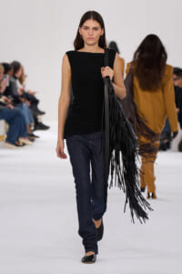 Model walking the runway in a black sleeveless ruched top, dark jeans, and a long black fringed bag over her shoulder; white studio backdrop.