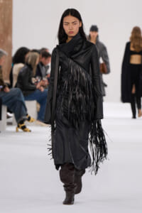Model walking a runway in a black fringe leather coat and knee-high boots at a fashion show.