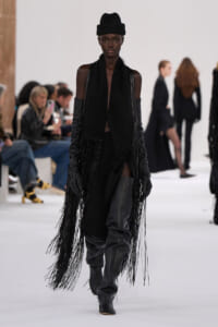 Runway model in all-black ensemble with long fringe coat, tall leather boots, and a black hat marching down the catwalk.