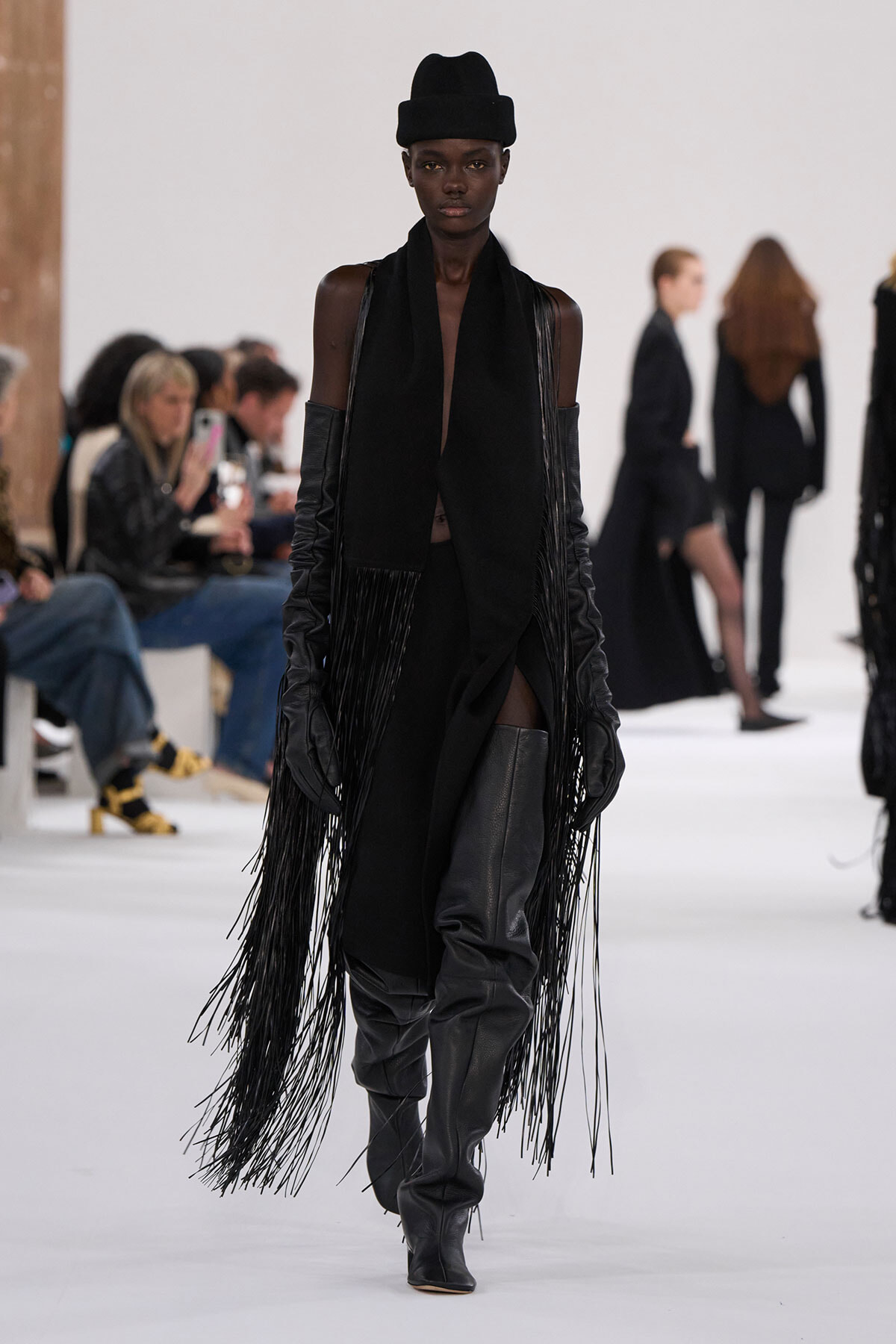 Runway model in all-black ensemble with long fringe coat, tall leather boots, and a black hat marching down the catwalk.