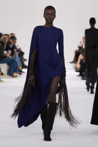 Model walks a cobalt blue draped dress with long fringe extensions, black gloves, and knee-high boots on a runway.