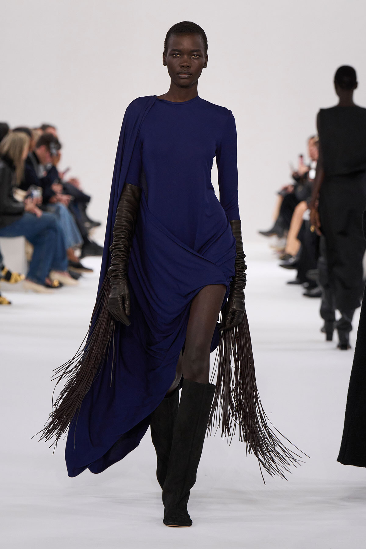 Model walks a cobalt blue draped dress with long fringe extensions, black gloves, and knee-high boots on a runway.