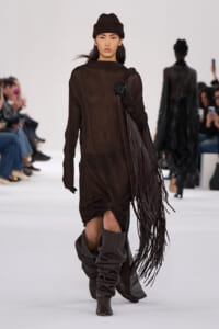 Model walking the runway in a dark brown ribbed dress with a long fringe bag and slouchy brown boots, wearing a matching brown beanie and black gloves.]'
