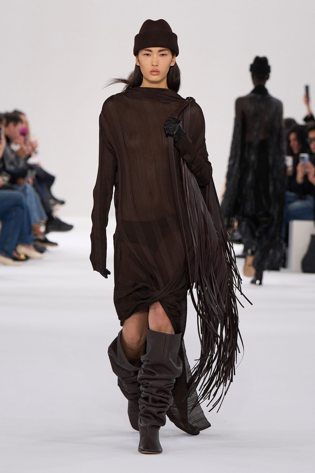 Model walking the runway in a dark brown ribbed dress with a long fringe bag and slouchy brown boots, wearing a matching brown beanie and black gloves.]'