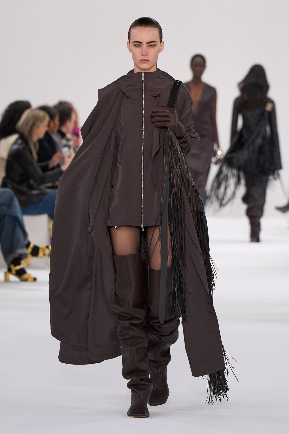 Model walking the runway in a dark brown cape-style coat, thigh-high boots, and fringe accessory with a long belt.