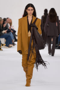 Model on a runway wearing a camel oversized suit with a black top, black gloves, and a large dark brown fringed leather bag over the shoulder.]','Runway fashion: camel suit with long fringe leather handbag slung over shoulder, audience in background.','Female model in a mustard camel suit with a black blouse, gloves, and a dark brown fringe tote.','Camel-colored suit with wide shoulders and matching boots, accentuated by a long leather fringe bag.','High-fashion runway look: camel suit, black shirt, leather glove, and a fringe handbag.