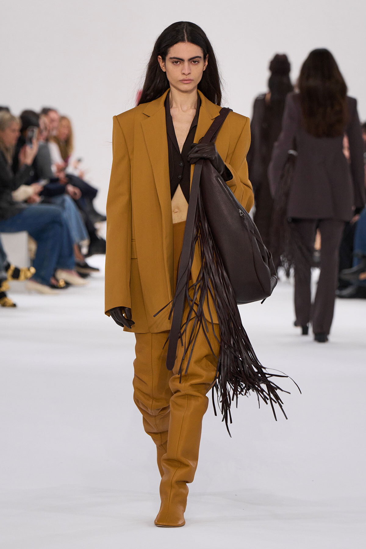 Model on a runway wearing a camel oversized suit with a black top, black gloves, and a large dark brown fringed leather bag over the shoulder.]','Runway fashion: camel suit with long fringe leather handbag slung over shoulder, audience in background.','Female model in a mustard camel suit with a black blouse, gloves, and a dark brown fringe tote.','Camel-colored suit with wide shoulders and matching boots, accentuated by a long leather fringe bag.','High-fashion runway look: camel suit, black shirt, leather glove, and a fringe handbag.
