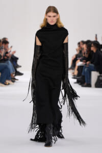 Model walks a white runway in a black sleeveless cape dress with long leather gloves and fringe tassels. - Fashion model in black turtleneck cape dress with fringe extensions and knee-high boots on a runway. - Black avant-garde outfit: cape-like shoulders, long fringe gloves, and fringed hem on a model. - Runway look featuring a black cape-dress, shoulder cutouts, and long dangling fringe at the sleeves. - Minimalist black ensemble with cape sleeves, tall boots, and fringe details, model walking.