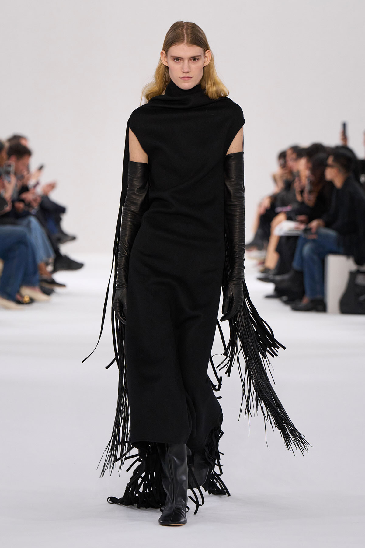 Model walks a white runway in a black sleeveless cape dress with long leather gloves and fringe tassels. - Fashion model in black turtleneck cape dress with fringe extensions and knee-high boots on a runway. - Black avant-garde outfit: cape-like shoulders, long fringe gloves, and fringed hem on a model. - Runway look featuring a black cape-dress, shoulder cutouts, and long dangling fringe at the sleeves. - Minimalist black ensemble with cape sleeves, tall boots, and fringe details, model walking.