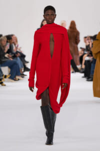 Model walks the runway in a bright red knit dress with a tall draped collar and a dramatic back opening, paired with black knee-high boots, audience in the background.