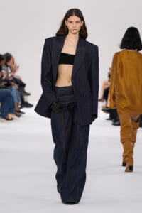 Model walking a fashion show in an oversized dark denim suit with a black bandeau top and black gloves.