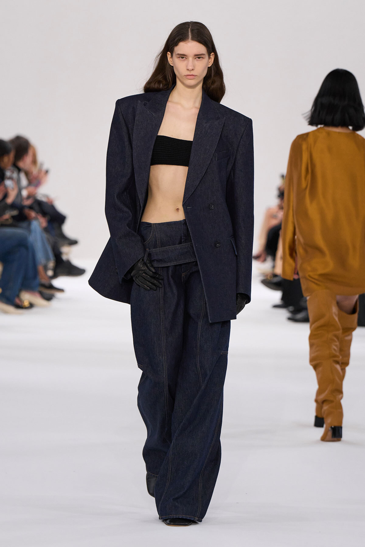Model walking a fashion show in an oversized dark denim suit with a black bandeau top and black gloves.