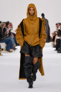 Model walks the runway in a tan oversized hooded coat with black shorts and tall black boots.