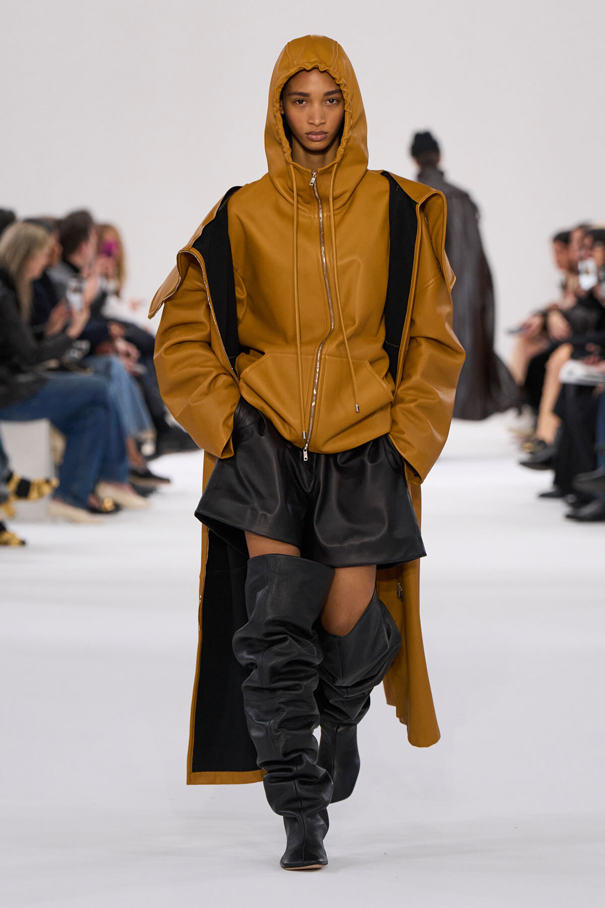 Model walks the runway in a tan oversized hooded coat with black shorts and tall black boots.