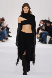 Model walks a fashion show in a black asymmetrical crop top, fringe cape, and thigh-high boots on a white runway.