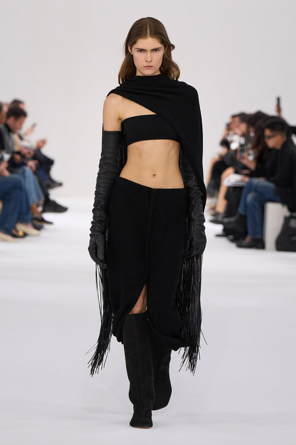 Model walks a fashion show in a black asymmetrical crop top, fringe cape, and thigh-high boots on a white runway.