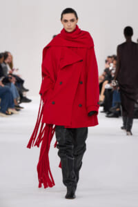Model walking a runway in a bright red oversized trench with fringe details and black leather pants under black boots.